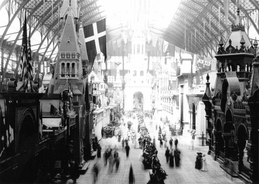 A black-and-white photograph features blurred human figures—as if in motion—in a wide aisle between rows of steepled buildings, one with a dark flag with a white cross on it.