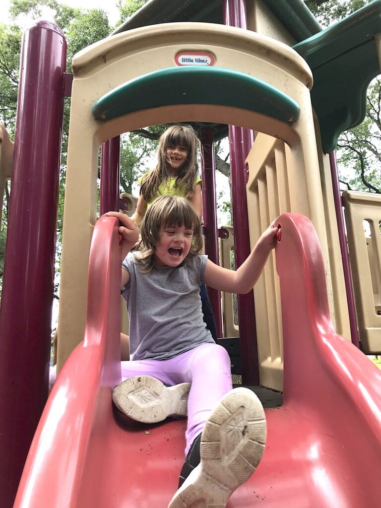 A young person with short brown hair in a gray T-shirt, pink leggings, and sneakers sits at the top of a red playground slide with a young person with longer brown hair in a yellow shirt standing behind them.