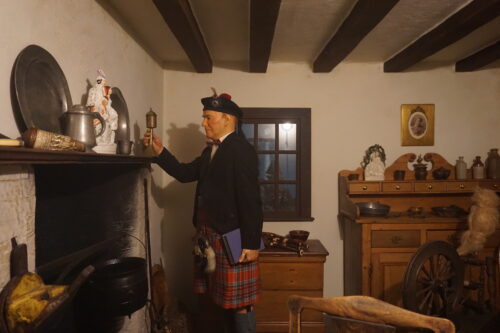 In a room featuring silver plates, wooden furniture with ceramics on top, and a black pot hanging in a fireplace, a wax figure of a person wearing a beret and red and green kilt holds a book with one hand and touches an object on the mantle with the other.