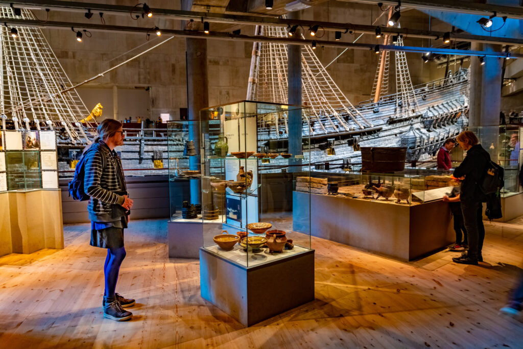 People stand in a room with wooden flooring; several tall, glass display cases with various objects and placards in them; and a large wooden ship installed in the background.