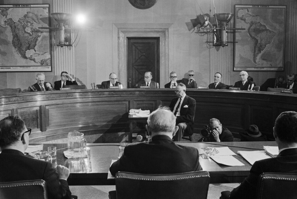 A black-and-white photograph features several people wearing glasses and black suits seated at a long, raised, curved wooden table. They are facing three other people in suits seated at a lower wooden table with several pieces of paper and a water pitcher on it.