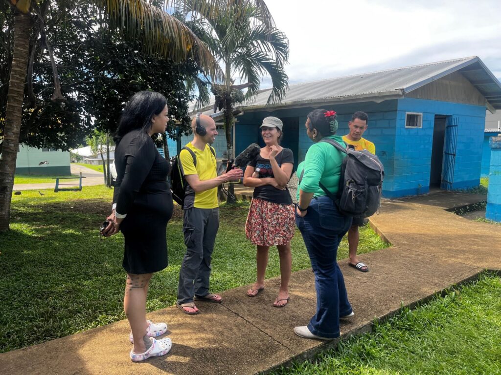 Five people stand in a semi-circle in an open, grassy area with a blue building in the background, conducting an audio interview.