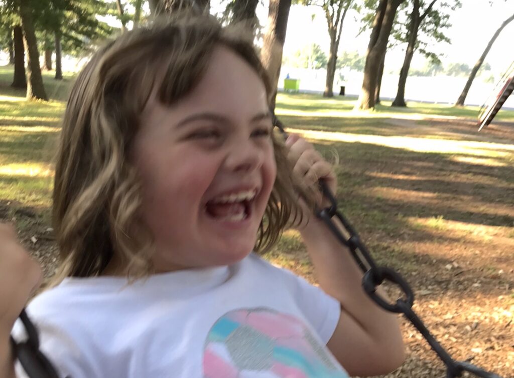 : A slightly motion-blurred image features a laughing girl wearing a white, pink, and blue T-shirt clutching the chain-link handles of a swing.