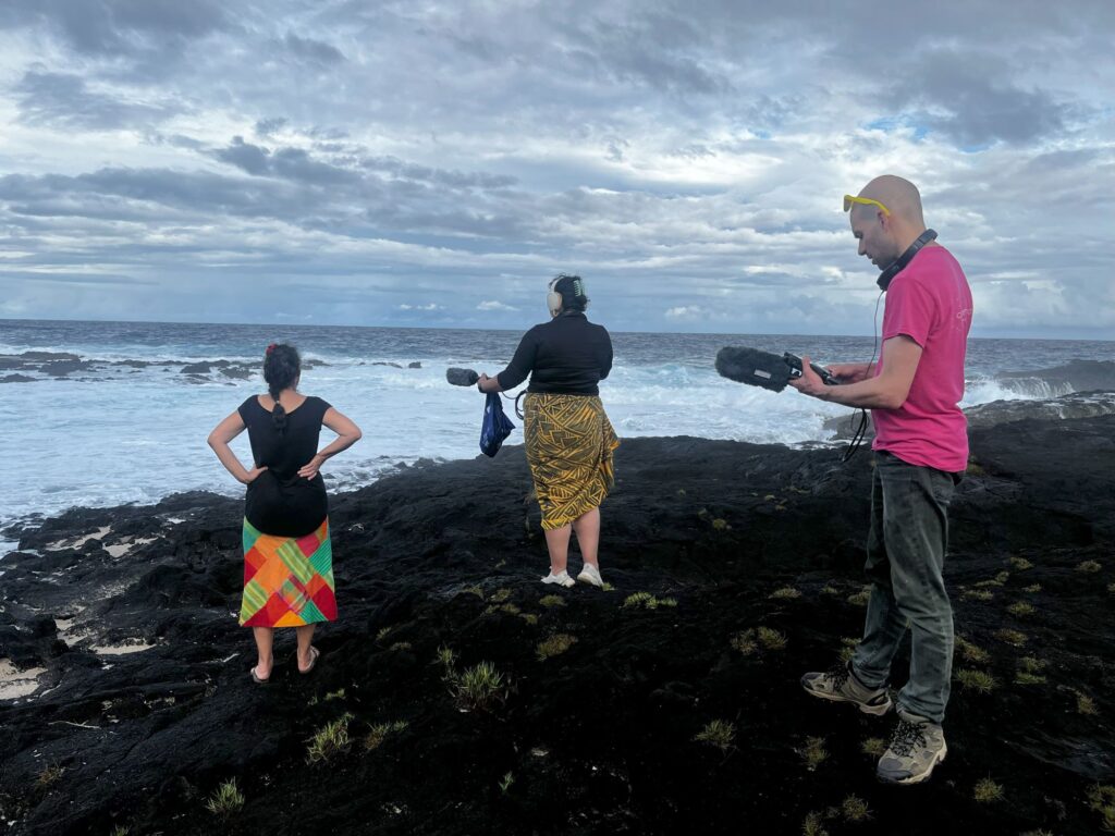 Three people stand on black lava next to the ocean conducting an audio recording.
