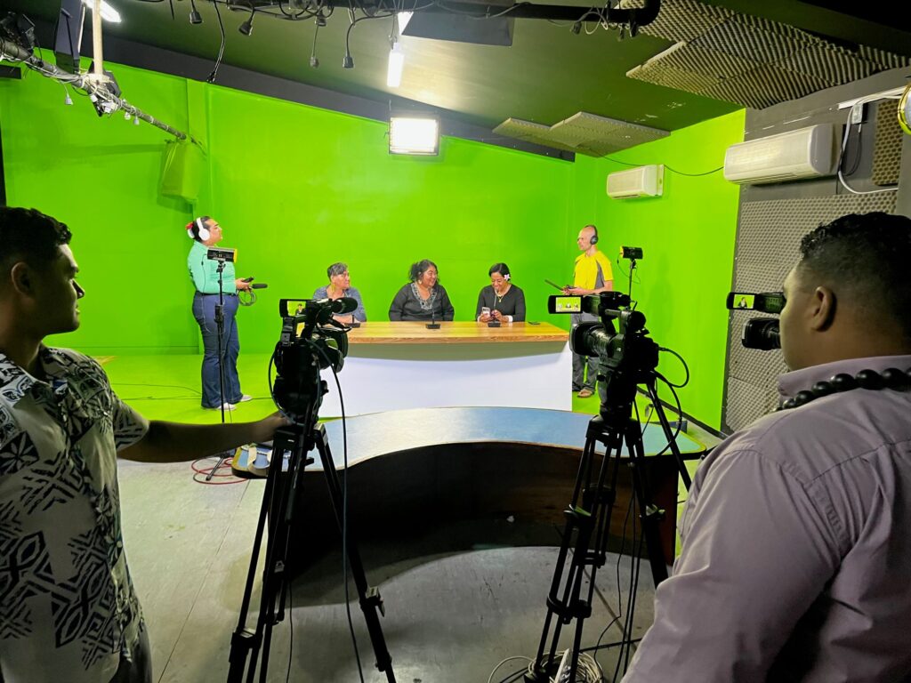 In a recording studio, three women sit at a desk with a bright green screen behind them, while a camera crew records them, and two audio engineers record them as well from the side.