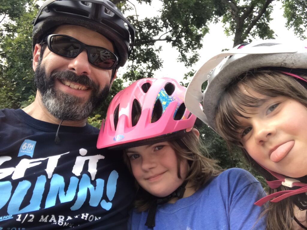 An adult with a salt-and-pepper beard and mustache stands beside two children. All three wear bike helmets—black, pink, and white, respectively. Tree branches are visible behind them.