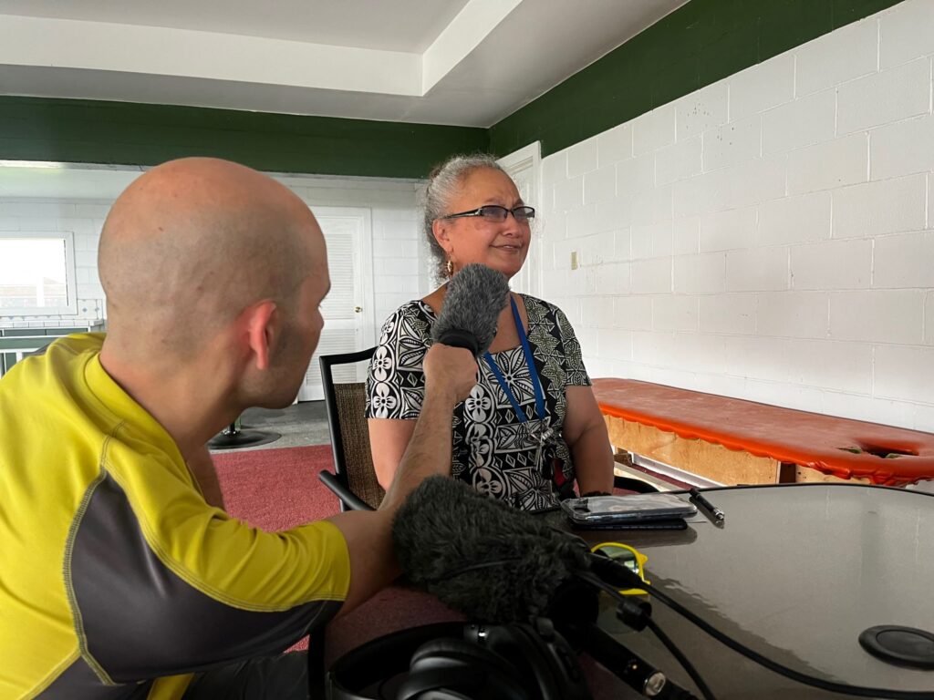 A man in a yellow shirt and bald heads leans in to interview a woman with grey hair and glasses.