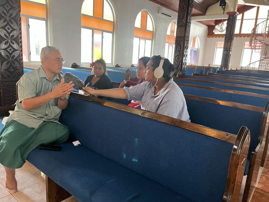 An older man is sitting in a church pew, gesticulating, while three women sit in the pew behind him. One woman is wearing earphones and leans in with a microphone to interview the man.