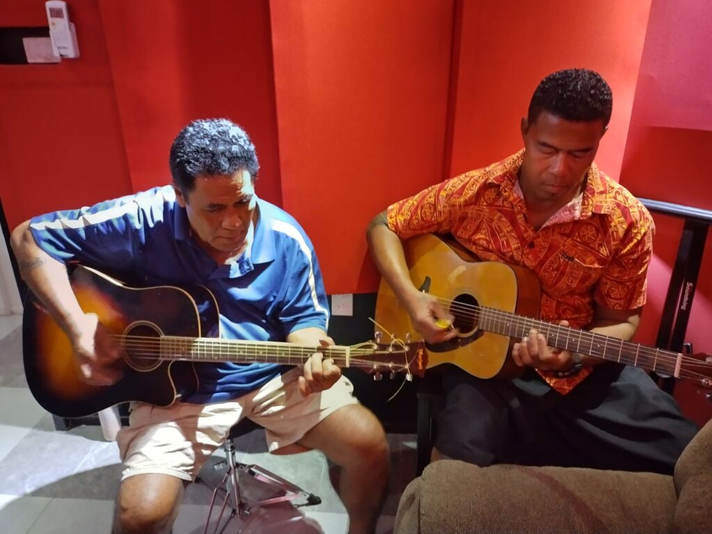 Two men sit in a recording studio with guitars on their knees.