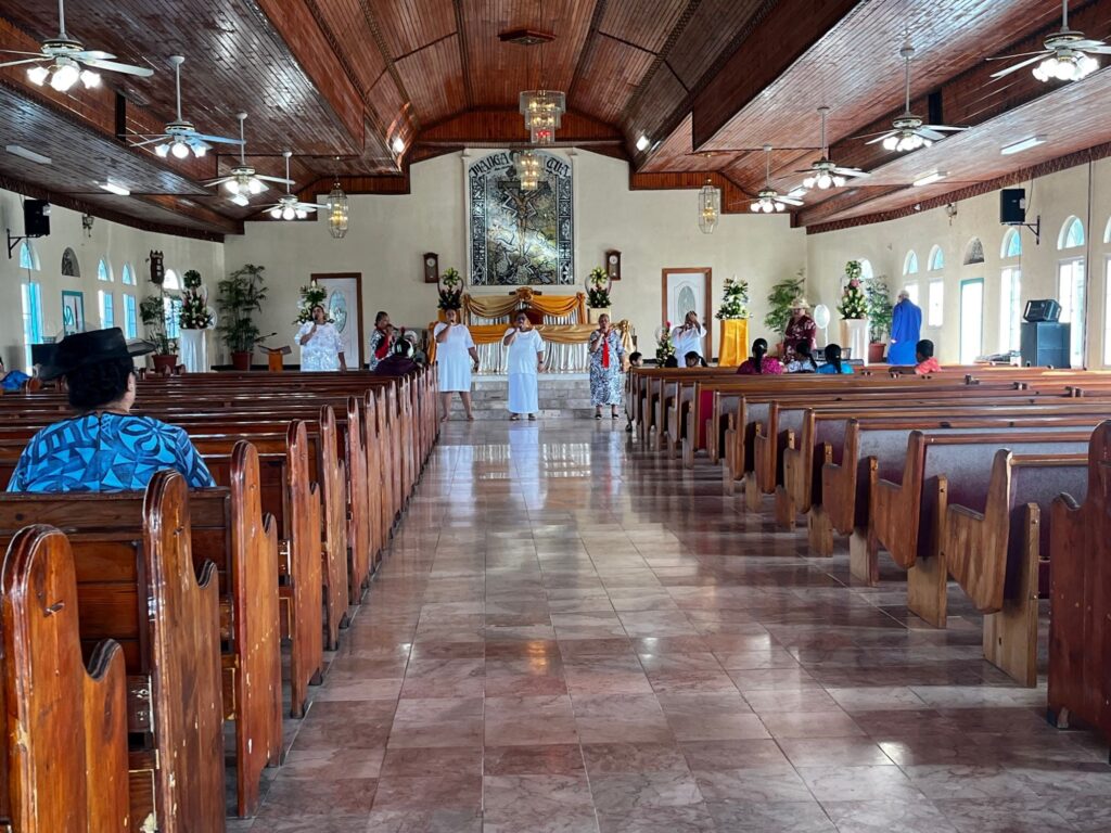 The interior of a church with wooden pews in neat rows, with singers in dresses standing in the front of the church by the altar.