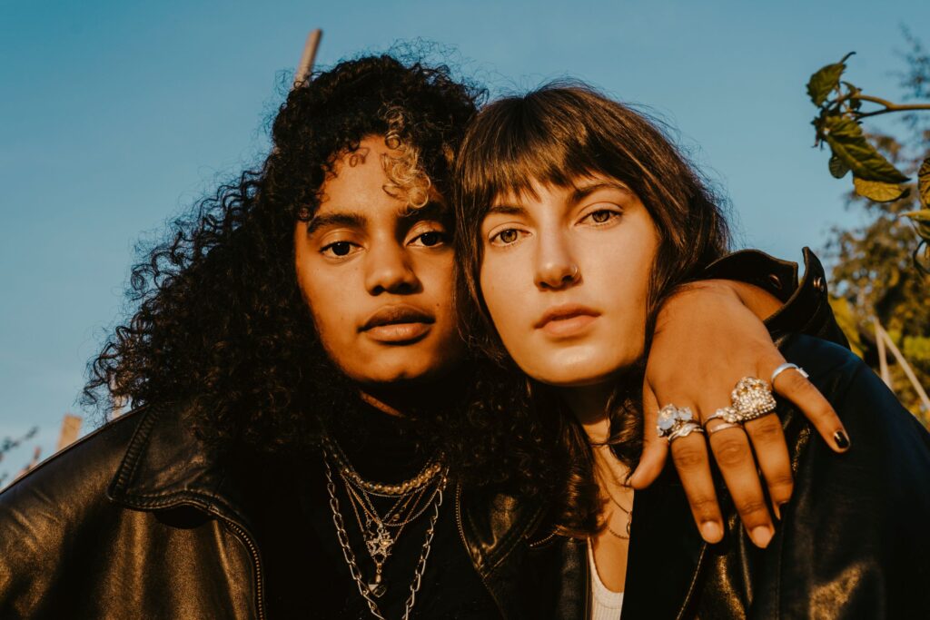 Portrait of two female friends with arm around outdoors on sunny day. One friend has dark curly hair and lots of jewelry, while the other girl has bangs and short cropped hair.