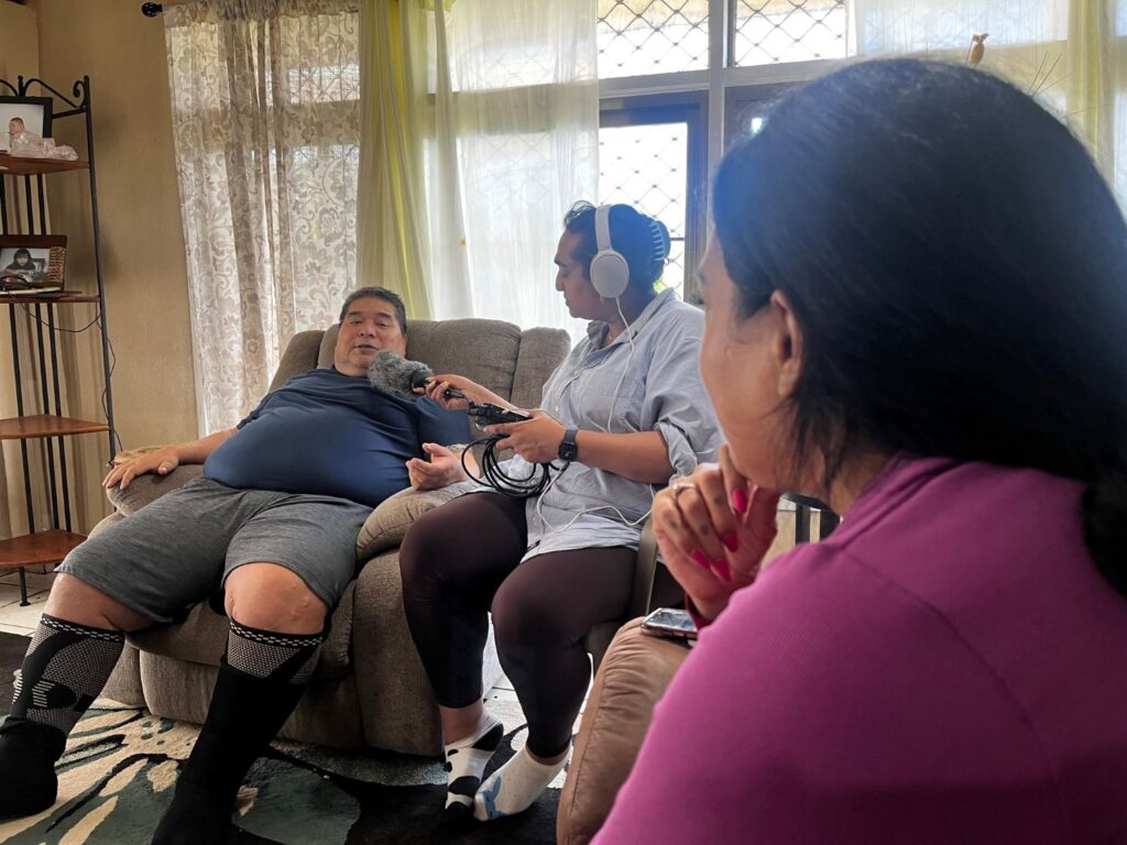 A heavy set man sits in a recliner being interviewed by two women.