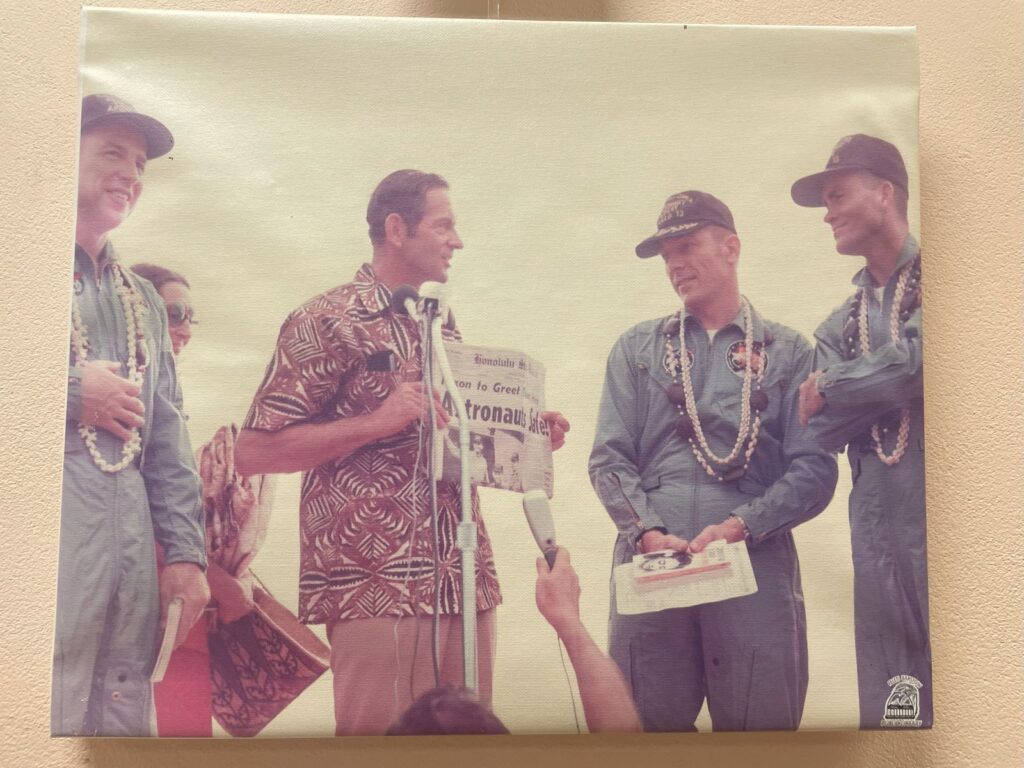 Four men stand on stage, three in astronaut jumpsuits. The fourth man is wearing an island style shirt and holding up a newspaper. One astronaut holds a book.