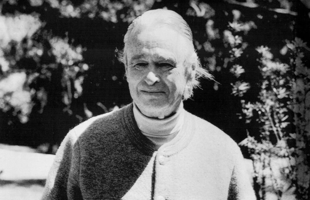A gray-haired man stands outside with his hands in the front pockets of his buttoned-up sweater coat.