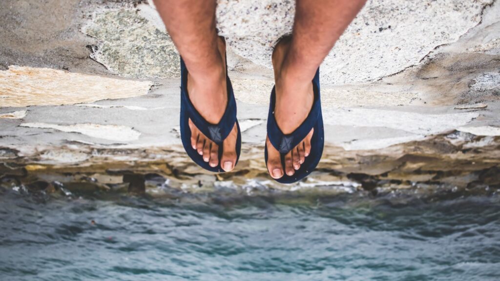 Two legs in flipflops hang off a cliff, taken from the perspective of the person sitting there. The ocean is soft focused in the distance. The overall effect is topsy-turvy.