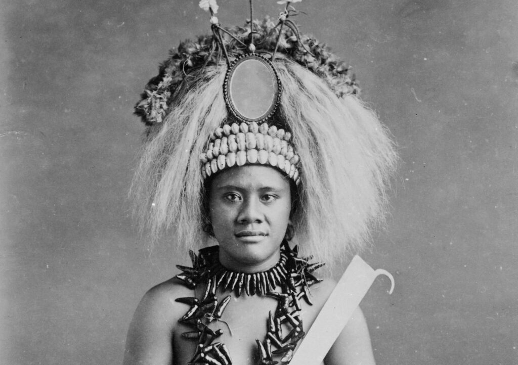A black and white photograph of a woman wearing an elaborate headdress and necklace, with bare shoulders exposed.