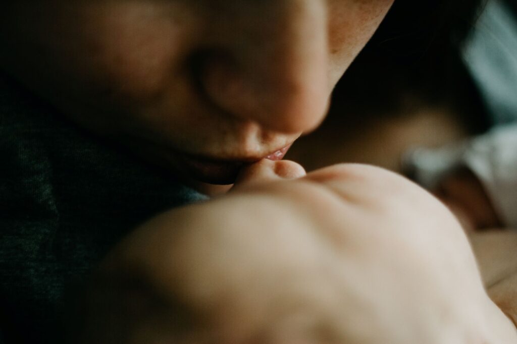In soft focus, a woman's lips gently kisses the tip of a baby's nose.