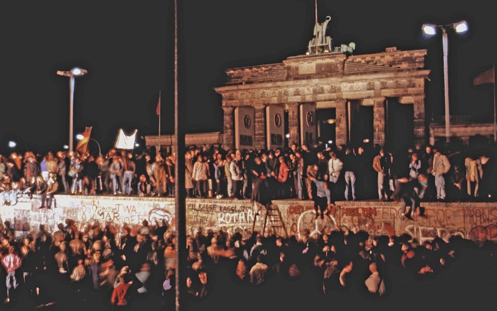 A large group of people stand by and sit on a long wall covered in graffiti, with some in the process of climbing it. Behind them looms a large stone pillared structure topped with a metal carving and a red flag.