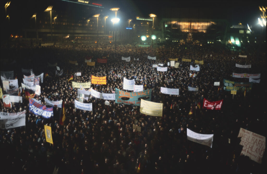 In a nighttime public square flanked by large buildings and lit only by streetlights, a large crowd of people gather. Many hold banners with writing on them.