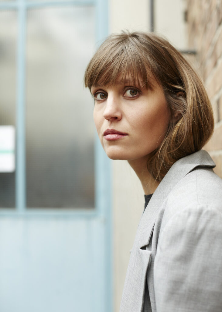 Visible from the bust up, a person with straight, shoulder-length brown hair wearing a gray suit jacket faces the left side of the image but turns to look straight at the viewer.