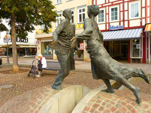 In a brick-covered public square surrounded by buildings, a metal statue features two people with flattened fronts and their hands extended sideways facing each other. They stand on opposite sides of a gap in the rounded brick hill they are built on.