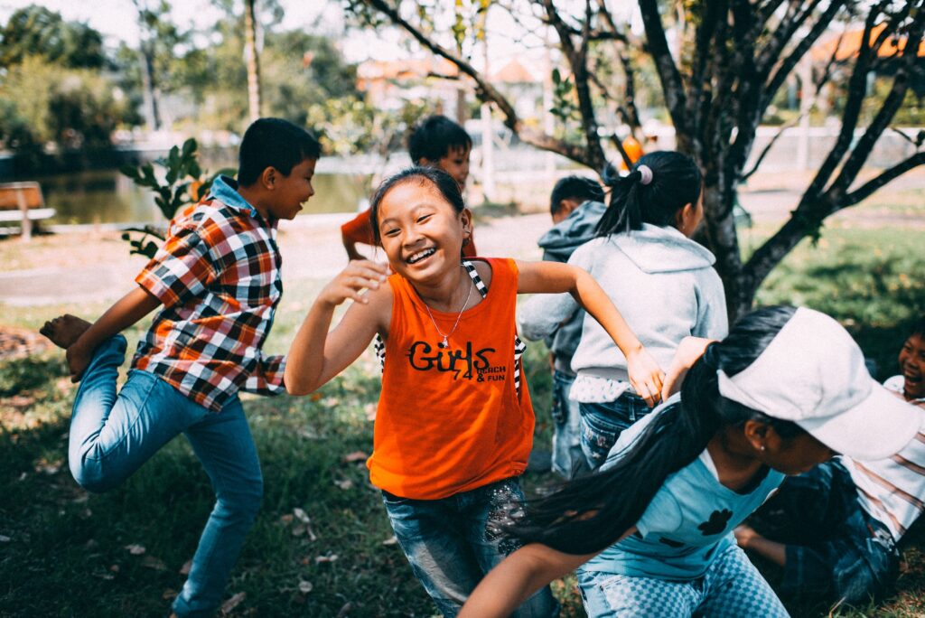 A small group of children run around under a tree, smiling happily.