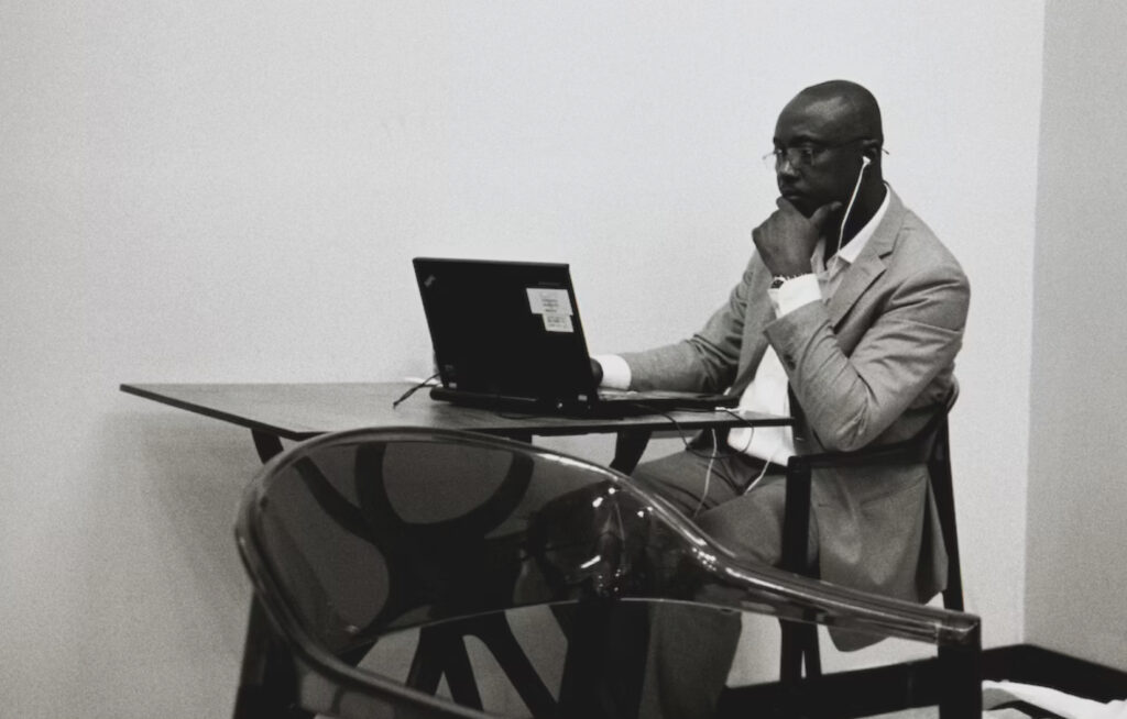 A Black man in a suit sits contemplatively before a laptop computer, perched on a table before him.