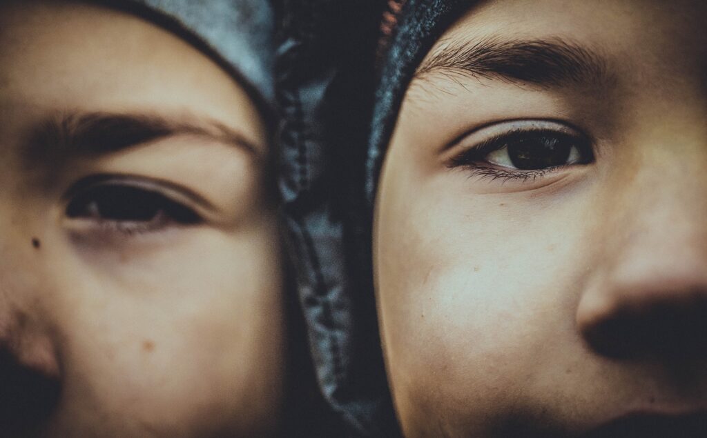 A pair of twins, two men, hold their heads close to one another, wearing winter hats.