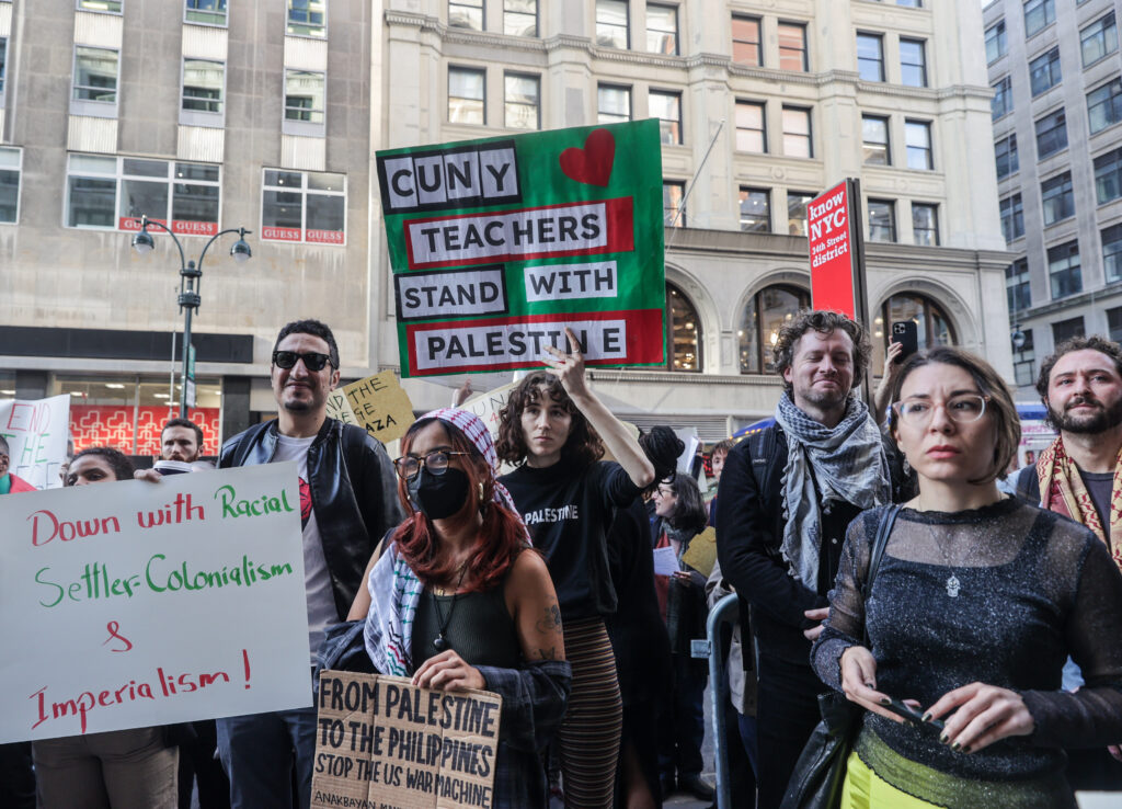 Several people stand in front of a large stone building holding posters with phrases including “CUNY Teachers Stand With Palestine,” “Down with racial settler-colonialism and imperialism,” and “from Palestine to the Philippines, stop the US war machine.”