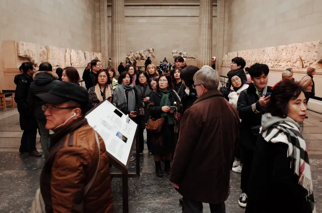 Several people crowd into the wide room of a museum exhibit. Beige statues depicting parts of posed human bodies line the side walls of the room.