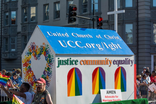 A crowd watches a parade float shaped like a building with rainbow-colored windows and a heart made of rainbow handprints. Text reads, “justice community equality” and words on a blue roof read, “United Church of Christ, UCC.org/lgbt.” A small sign propped up against the float reads, “Amen. Advocate for Marriage Equality.”