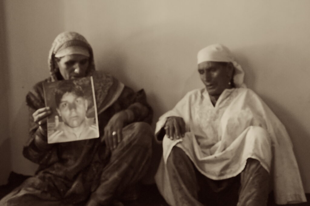 In sepia tones, a slightly blurry image features two people wearing head coverings and tunics seated against a blank wall. The one on the left holds up a piece of paper with a person’s face printed on it.