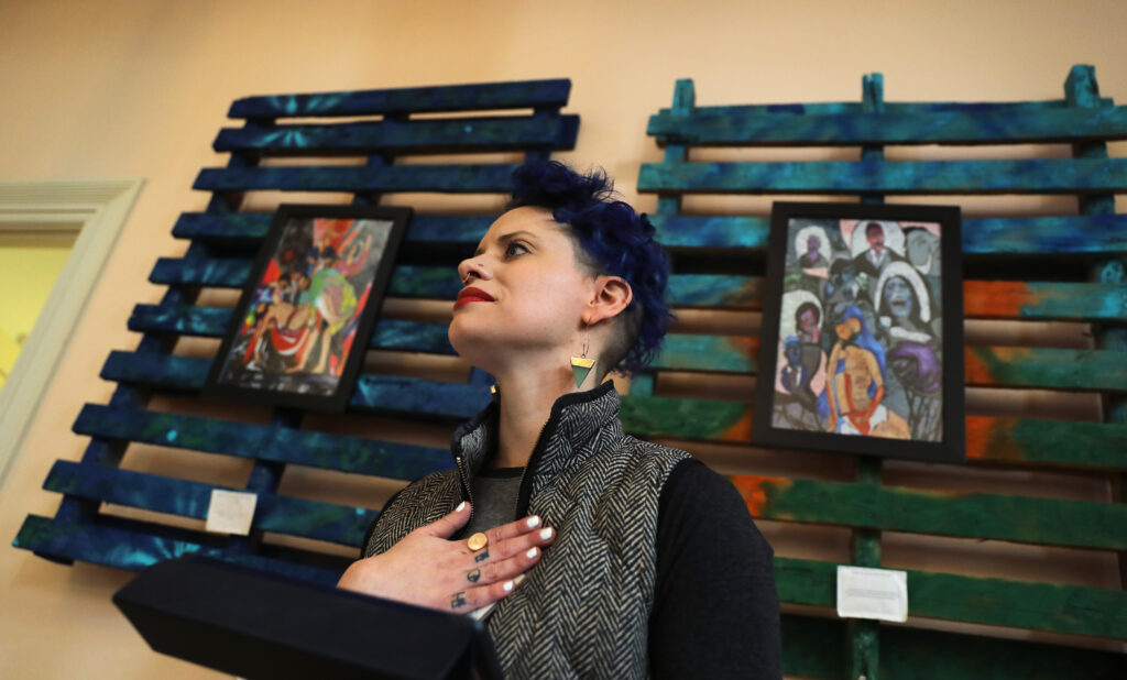 A person with short, dark-blue hair wearing triangle earrings and a gray and black jacket stands with their right hand over their heart. Behind them, two framed paintings hang on green wooden structures.