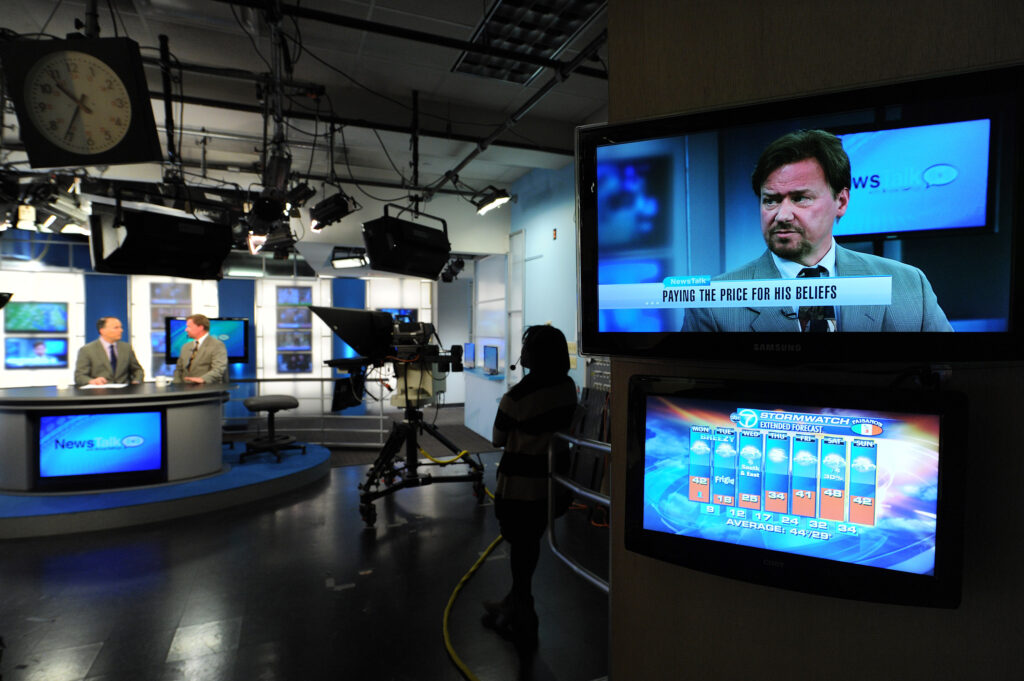 Two people wearing suits and ties sit at a newsroom table. In the foreground, a screen features the close-up image of one of the seated people overlaid with text that reads, “paying the price for his beliefs.”