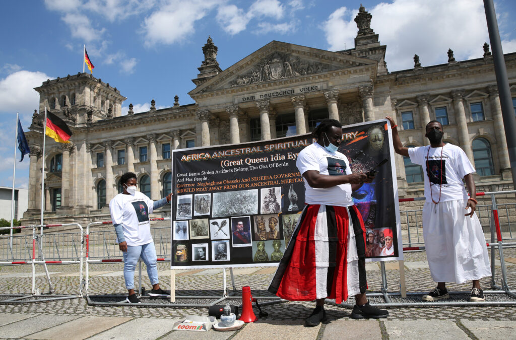 Tres personas de piel oscura llevan camisetas blancas con imágenes cuadradas. Dos sostienen un gran cartel en inglés que dice: “Africa Ancestral Spiritual Ministry.” El texto de abajo dice: "Great Queen Idia Eyes. Great Benin Stolen Artifacts Belong the Oba of Benin and His People. Governor Nogeghase Obaseki and Nigeria government should stay away from our Ancestral Heritage". Visible detrás del titular, una colorida máscara llena la parte derecha del cartel. Debajo hay varias imágenes de archivo de objetos, grupos de personas africanas y diferentes hombres europeos.