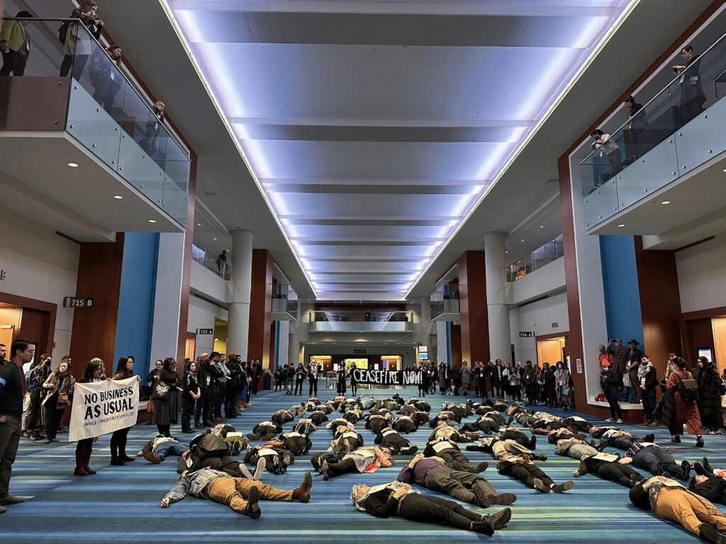 In a building’s large, carpeted atrium, several people in button-down shirts, khakis, and slacks lie on the floor. People on the perimeter hold up two signs, one that reads, “No Business as Usual While Canada Funds Genocide” and another that says, “Ceasefire Now!”