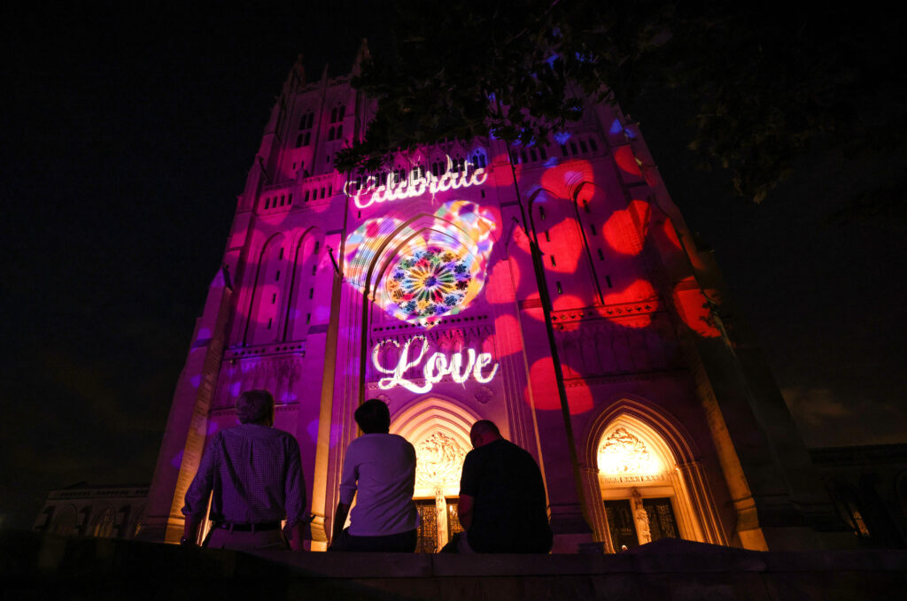 People look up at an illuminated cathedral whose entrance glows orange against a black sky. A light projection of multicolored hearts shines on the center of its facade along with text that reads, “Celebrate Love.”