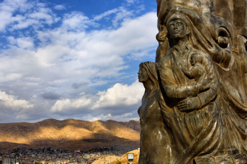 A large, brown sculpture depicts a cluster of people looking outward, one cradling a child in their arms. A village and blue sky are visible in a distant horizon.