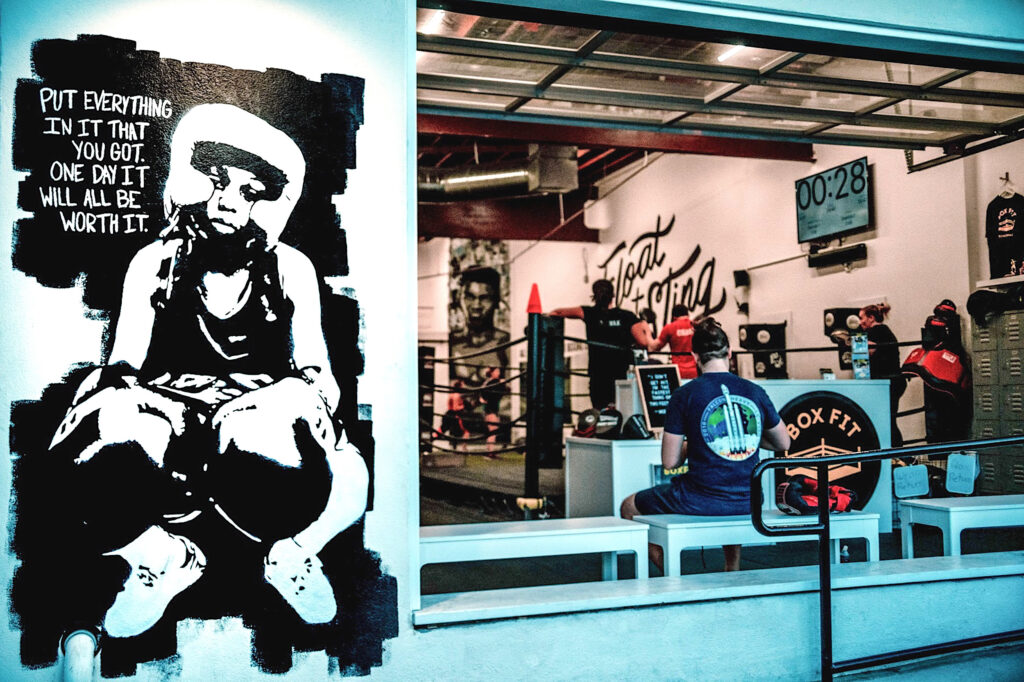 Looking in from a gym entryway, a photograph features a black-and-white mural on the front wall that depicts a person wearing a headguard and boxing gloves under text that reads, “Put everything in it that you got. One day it will all be worth it.” To the right, people are using a boxing ring behind a podium with a black circular plaque that reads, “BoxFit.”