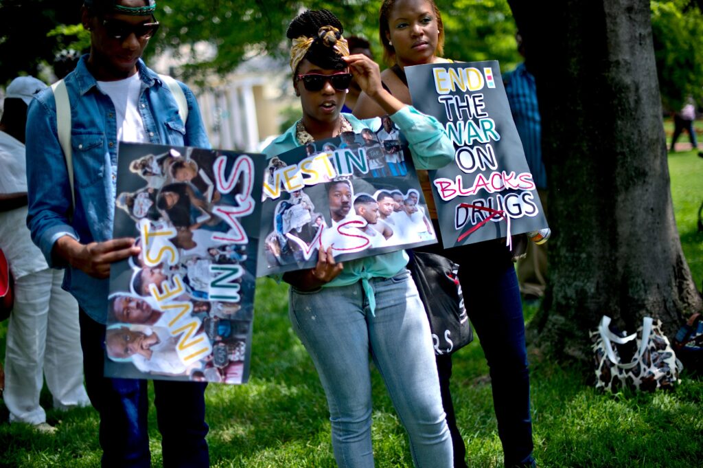 Three people stand on a grassy field holding signs with pictures of young people’s faces pasted on them. Also on the signs, in colorful text, are two phrases: “Invest in Us” and “End the War on Drugs” with the word “Drugs” crossed out and the word “Blacks” added before it.