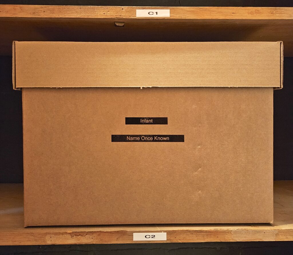 A large, tan cardboard box sits on a shelf with a label that reads “C2” below it. On two small pieces of black tape on the box’s front side, white letters read “Infant” and “Name Once Known.”