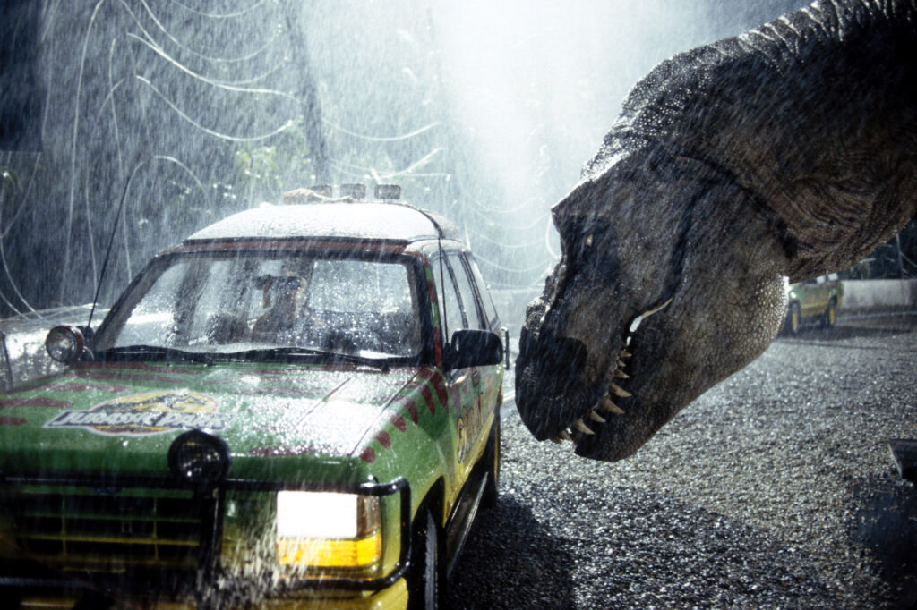In a dark and rainy forest, a large Tyrannosaurus rex stares into a green car with its headlights on. The car has a yellow and black graphic on its hood with text that reads, “Jurassic Park.”