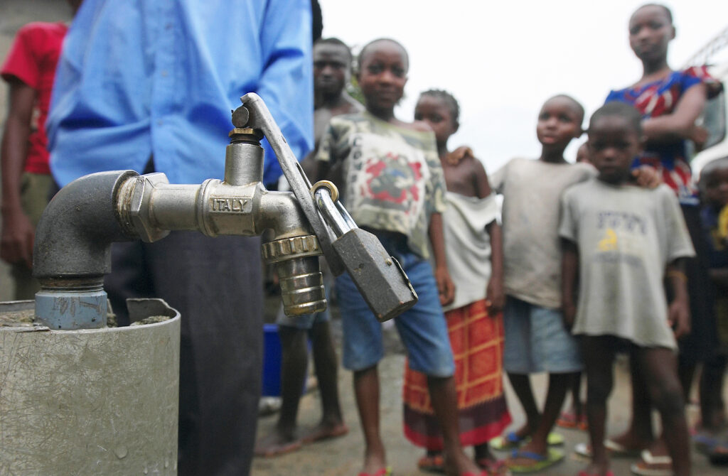 A crowd of adults and children wearing T-shirts, flip-flops, and denim shorts or sarongs looks at a water tap with the word “Italy” stamped on its side and a giant silver lock on it.