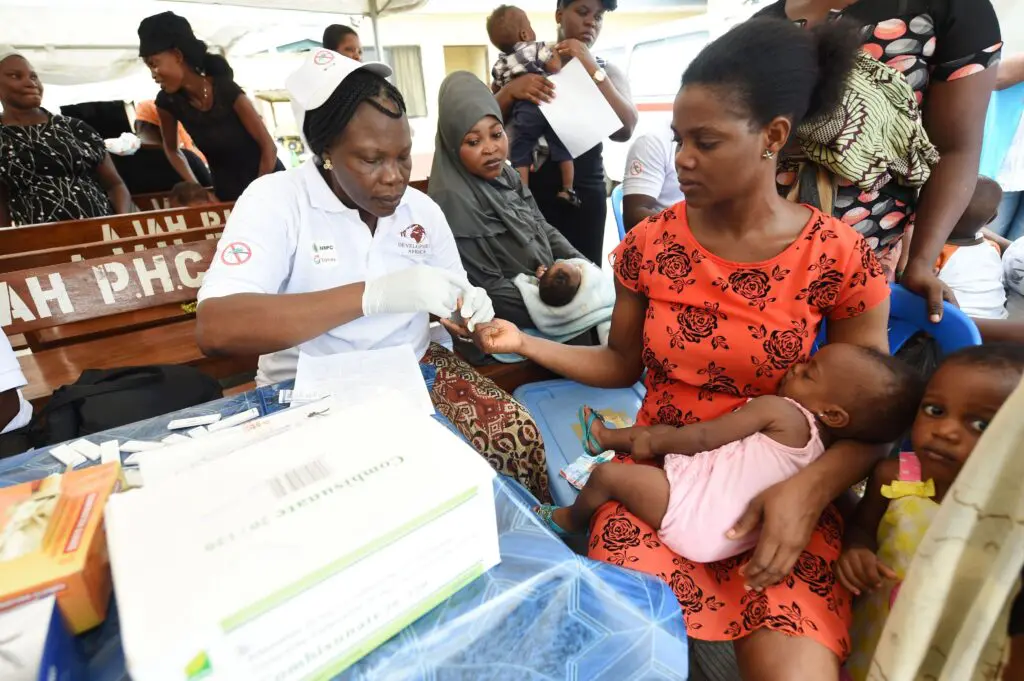 Surrounded by an otherwise occupied crowd, a person wearing a white cap and shirt uses gloved hands to work with the finger of a person seated beside them. The second person wears a red-orange dress and holds a baby while a slightly older child stands beside them, looking at the viewer.