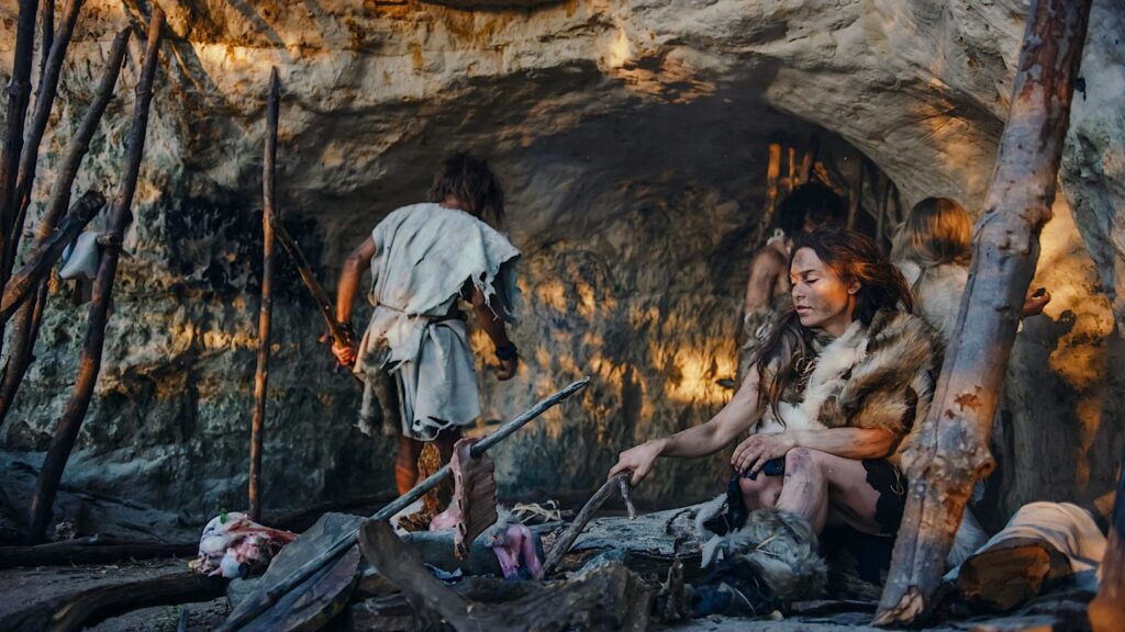 A computer-rendered graphic depicts a group of people in torn clothes and furs with black smudges on their faces and bodies. They gather in front of a rock overhang, with one sitting in front and poking at a pile of wood near which are dangling pieces of meat.