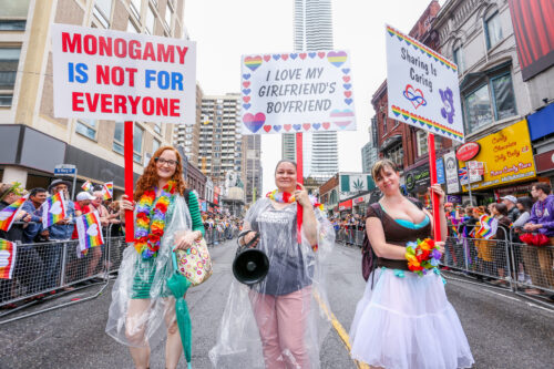 On a paved city street fenced off and lined with people, three people wearing colorful clothing stand and hold signs. These read: “Monogamy is not for everyone,” “I love my girlfriend’s boyfriend,” and “Sharing is Caring.”