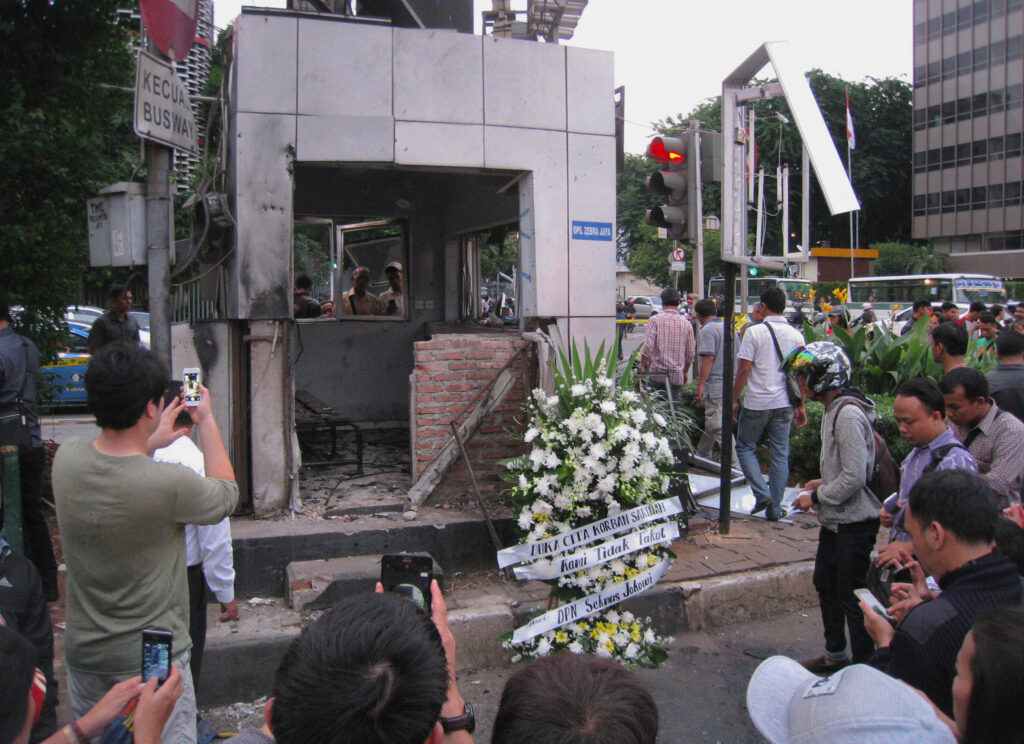 A crowd of people, with several taking pictures on cellphones, surround a damaged structure in a public square. The small building, with its tiling cracked and wooden seating splintered, has a large arrangement of white flowers in front of it.