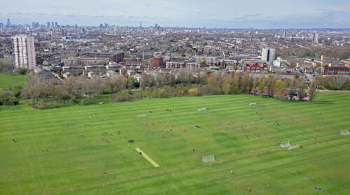 A zoomed-out photograph shows a large green, grassy field with several football pitches in front of a line of trees. The trees separate the field from a large cityscape in the background.