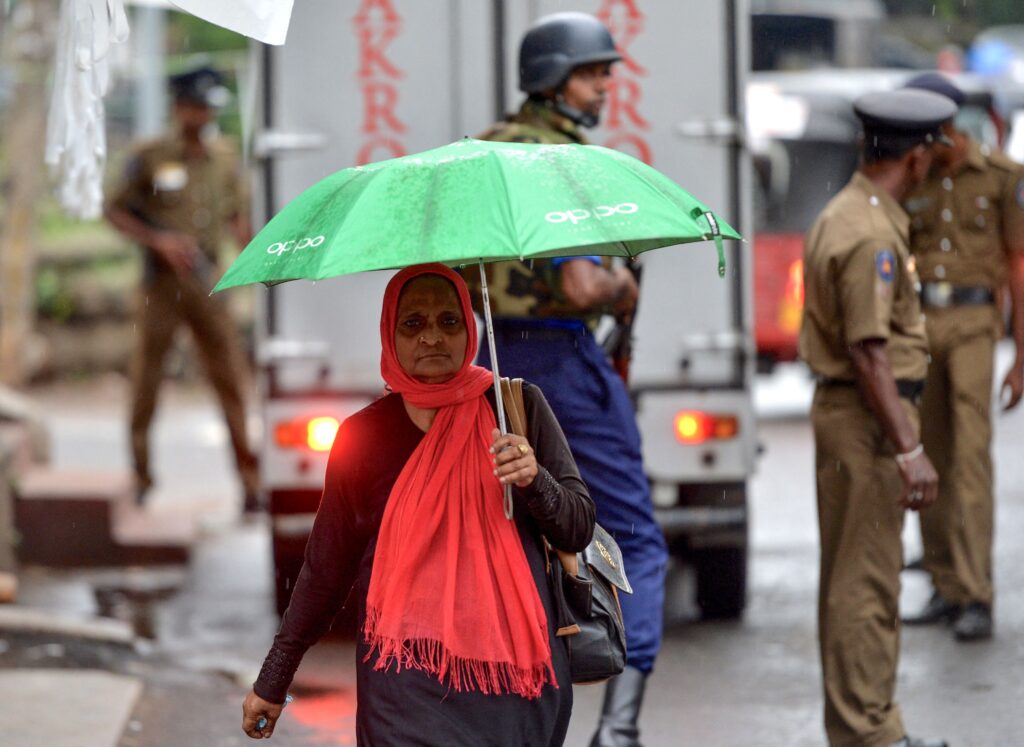 A person wearing a long-sleeved black maxi dress and a red headscarf holding a green umbrella walks on a public street with a silver truck and several people in black hats, helmets, and khaki uniforms in the background.