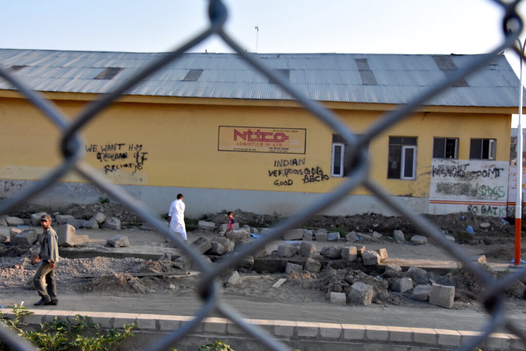 Taken through a wire fence, a photo shows a few people passing by a yellow building with several works of graffiti written on its wall and piles of square stone blocks in front of it.